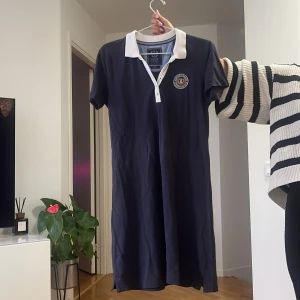 Marinblå kortklänning från Gant - Sportig marinblå kortklänning från Gant med vit krage och knappslå. Klänningen har korta ärmar och en broderad logga på bröstet. Perfekt för en preppy look och tillverkad i mjuk bomull för maximal komfort.