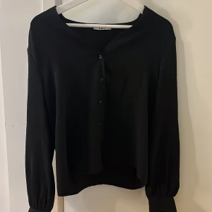 Svart blus med knappar från Nelly - Stilren svart blus i storlek XS. Blusen har v-ringning, knappar framtill och långa ärmar med lätt puff vid ärmslutet.  Materialet känns lätt och luftigt. Bara använd 1 gång, hör av dig vid frågor💗💗💗