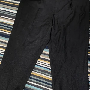 Svarta kostymbyxor från Taylor's - Klassiska svarta kostymbyxor från Taylor's Trouser Collection. Byxorna är tillverkade i en blandning av 60% ull och 40% polyester, har rak passform och stängs med knapp i midjan. Perfekta för en clean och stilren look. Midjan är 112 cm och längden är 102 cm