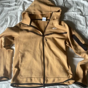 Nike hoodie från Nike med dragkedja - Säljer en Nike hoodie från Nike med hel dragkedja och huva. Tröjan har svarta detaljer längs dragkedjorna och en extra ficka med dragkedja på ena ärmen. Perfekt för en chill och sportig stil.