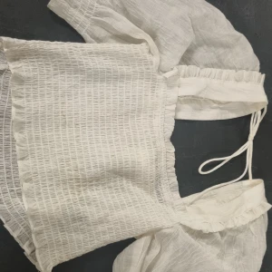 Vit blus med puffärmar från H&M - Supersöt vit blus från H&M med puffiga ärmar och smockad, croppad passform. Toppen har knytband i ryggen och är tillverkad i ett lätt, luftigt bomullstyg med struktur. Perfekt för en trendig och romantisk look.