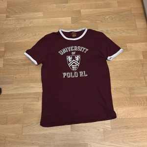Vinröd t-shirt Polo Ralph Lauren - Vinröd t-shirt från Polo Ralph Lauren med vit text och emblem på bröstet. T-shirten har vita kontrastkanter vid hals och ärmslut, klassisk rund hals och korta ärmar. Slim fit-modell i mjuk bomull, perfekt för en avslappnad och snygg stil.