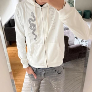 Vit hoodie från Trapper med strassorm - Säljer en vit hoodie från Trapper med dragkedja, huva och kängurufickor. På bröstet finns en svart orm i strass och Trapper-loggan i silvriga stenar. Mjuk bomullskänsla och snygga detaljer som dragsko i huvan.