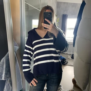 Randig stickad tröja med v-ringning - Mörkblå och vit randig stickad tröja med v-ringning och krage. Tröjan har långa ärmar, avslappnad passform och breda ribbade muddar. Perfekt för dig som gillar en chill och stilren look.