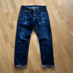 Selvedge jeans från Nudie Jeans - Snygga mörkblå jeans från Nudie Jeans med klassisk femficksdesign och kontrastsömmar. Jeansen har raka ben och slitningar på både baksida och vid benslut. Tillverkade i selvedge denim så du kan lita på att de håller länge. Mått = 102 cm längd, 41 cm midja