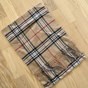 Beige rutig halsduk med fransar - Stilren beige halsduk med klassiskt rutigt mönster i svart, vitt och rött. Halsduken har snygga fransar i ändarna och passar perfekt för att lyfta din outfit under kyliga dagar. Mjuk och skön att bära. Oanvänd 