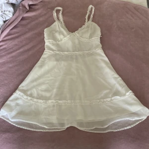 Vit omlottklänning från H&M Divided - Supersöt vit kortklänning från H&M Divided med volangdetaljer och omlottknytning i ryggen. Klänningen har smala axelband och är tillverkad i ett lätt, transparent material som ger en luftig känsla. Perfekt för sommarens varma dagar.