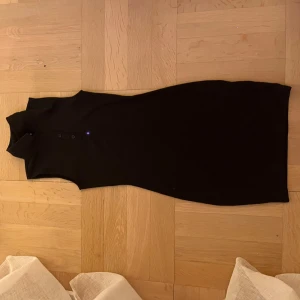 Svart ärmlös ribbad klänning - Svart, ärmlös klänning med ribbad struktur och hög krage. Klänningen har knappar framtill vid halsen och en tight passform som framhäver siluetten. Perfekt för dig som gillar minimalistisk och stilren look.