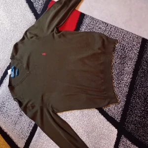 Olivgrön tröja från Polo Ralph Lauren - Olivgrön långärmad tröja från Polo Ralph Lauren med klassisk v-ringning och röd broderad logga på bröstet. Tillverkad i 100% bomull, perfekt för dig som gillar stilren och enkel design med en touch av preppy.