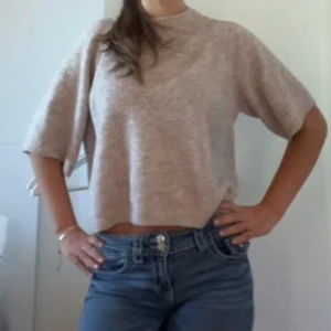 Beige stickad croppad tröja  - En beige stickad topp med croppad passform och halvlånga ärmar. Toppen har en enkel och stilren design med hög hals och mjukt material som känns skönt mot huden. Perfekt för dig som gillar en avslappnad men trendig look.