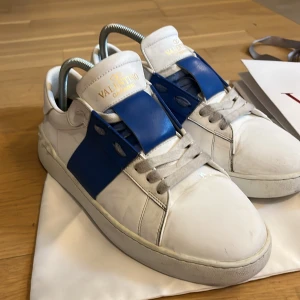 Valentino Garavani vita sneakers med blå detalj - Säljer ett par vita Valentino Garavani sneakers i skinn med blå bred rem över vristen och ikoniska nitar på hälen. Skorna har snörning och rund tå, och kommer med originalkartong och dustbag. Perfekta för dig som gillar lyxiga och trendiga sneakers.