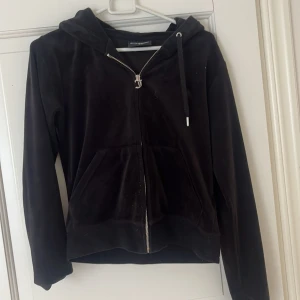 Svart velour hoodie Juicy Couture S - Mysig svart hoodie från Juicy Couture i mjuk velour med dragkedja framtill, huva och snörning. Klassisk modell med kängurufickor och långa ärmar. Perfekt för en chill och trendig look.