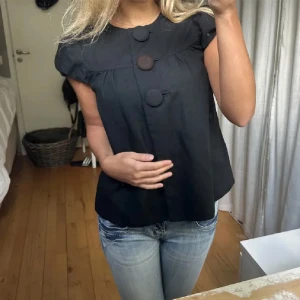Svart blus med stora knappar - Supersöt svart blus från H&M med korta puffärmar och tre stora tygklädda knappar framtill. Blusen har en lös passform och rund halsringning, perfekt att styla med jeans eller kjol. Materialet känns som en mjuk skjorta och har ett lätt glansigt utseende.