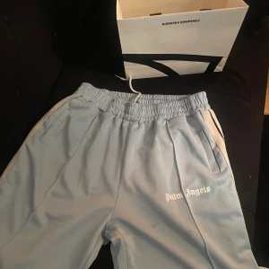 Palm Angels ljusblå mjukisbyxor - Ljusblå mjukisbyxor från Palm Angels med vita sidoränder och broderad logga på vänster ben. Byxorna har elastisk midja med dragsko och raka ben. Perfekta för en chill och sportig look.