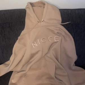 Beige hoodie från NICCE - Säljer en beige hoodie från NICCE med broderad logga framtill. Tröjan har huva med snörning och en stor magficka. Perfekt för dig som gillar en clean och chill stil.