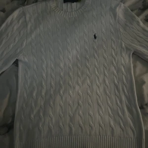Vit kabelstickad tröja Polo Ralph Lauren - Kabelstickad vit tröja från Polo Ralph Lauren med klassisk rund halsringning och broderad logga på bröstet. Tröjan har långa ärmar och ribbade muddar vid ärmslut och nederkant. Perfekt för en clean och stilren look.