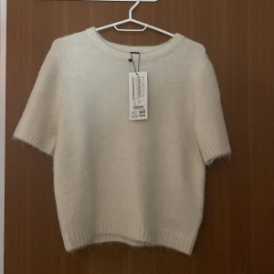 Vit stickad kortärmad tröja från H&M - Supermjuk vit stickad tröja från H&M med korta ärmar och rund halsringning. Tröjan har en fluffig känsla och ribbade kanter vid ärmslut och nederkant. Perfekt för lager-på-lager och en clean look.