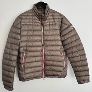Beige Moncler dunjacka strl 4 - Snygg beige dunjacka från Moncler med horisontella sömmar, hög krage och klassisk logga på ärmen. Jackan har två fickor med dragkedja och rödvit randig detalj vid dragkedjorna. Perfekt för kyliga dagar och riktigt lätt att bära. Storlek 4 (S-M)