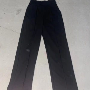 Svarta kostymbyxor med raka ben - Säljer ett par svarta kostymbyxor med hög midja och raka, vida ben. Byxorna har pressveck framtill och sidfickor. Perfekta för en clean och stilren look. Materialet känns mjukt och har en snygg fall.