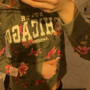Grön blommig hoodie CHICAGO - Säljer en grön hoodie från Awesome Days med stort vitt CHICAGO-tryck på bröstet. Tröjan har blommigt mönster i rosa och röda toner, huva och magfika. 