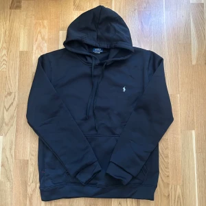 Svart hoodie från Polo Ralph Lauren - Svart hoodie från Polo Ralph Lauren med klassisk huva och snörning. Tröjan har en broderad logga på bröstet och en stor magficka framtill. Tillverkad i mjuk bomull och har långa ärmar med muddar. Perfekt för en chill och stilren look.
