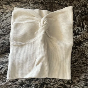 Vit ribbad stickad topp med knut - Superfin vit stickad topp med ribbad struktur och snygg knutdetalj framtill i bröstet. Toppen är figurnära och har en mjuk, stretchig känsla som gör den bekväm att bära. Perfekt för dig som gillar en clean och trendig look.
