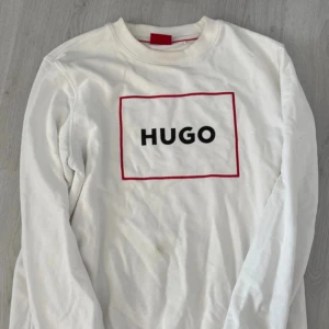 Vit sweatshirt från HUGO med röd detalj - Vit långärmad sweatshirt från HUGO med svart logotyp på bröstet och en röd rektangulär ram runt trycket. Tröjan har rund hals och ribbade muddar. Materialet är mjuk bomull som känns skön mot huden. Perfekt för en clean och stilren look. De små fläckarna går att få bort i tvätten 