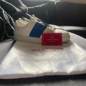 Valentino Garavani Open sneakers vit/blå - Snygga Valentino Garavani Open sneakers i vitt skinn med bred blå rem över vristen. Klassisk rund tå, vita snören och diskret logga på plösen. Kommer med originalpåse och kort. Perfekta för dig som vill ha en stilren men ändå unik look.OBS INGEN BOX!