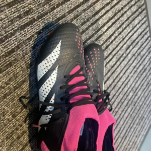 Svarta och rosa fotbollsskor med snörning - Säljer ett par svarta fotbollsskor med vita detaljer och rosa inslag. Skorna har snörning, platt sula med dobbar och en högre strumpliknande kant i rosa. Perfekta för dig som vill sticka ut på planen med snygg design och bra grepp.