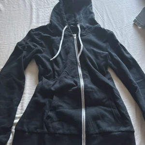 Svart hoodie med dragkedja och huva - Svart hoodie med vit dragkedja och snörning i huvan. Tröjan har långa ärmar och en klassisk känguruficka framtill. Perfekt för chill dagar eller när du vill ha en enkel och stilren look.