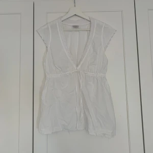 Vit blus från Esprit - Söt vit topp från Esprit med spetsdetaljer vid ärmar och halsringning. Djup v-ringning framtill, knytband i midjan och ärmlös design med broderade detaljer. 
