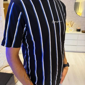 Svart randig t-shirt från Mannen - Svart t-shirt med vita och blå vertikala ränder från Mannen. Klassisk passform med korta ärmar och rund hals. Tunt och mjukt material, perfekt för en chill look. Liten vit logga broderad på bröstet. Passar bra S/M/L storlek.