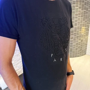 Svart Kenzo t-shirt med tigertryck - Svart t-shirt från Kenzo med ikoniskt broderat tigerhuvud och texten 'Kenzo Paris' på bröstet. Klassisk passform och rund halsringning. Tillverkad i mjuk bomull som känns skön mot huden. Perfekt för dig som vill ha en stilren men ändå statement t-shirt. Passar vra som oversize modell.