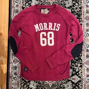 Röd stickad tröja Morris - Vinröd stickad tröja från Morris Gents med vita bokstäver och siffran 68 framtill. Tröjan har svarta förstärkningar på armbågarna, ribbade muddar och rund hals. Perfekt för dig som gillar sportig och klassisk stil.