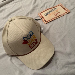 Beige keps från Dsquared2 Summer Lovers - Snygg beige keps från Dsquared2 med färgglad broderad text framtill och röd kant runt skärmen. Nästan slutsåld överallt och nypris runt 2700kr, pris kan diskuteras