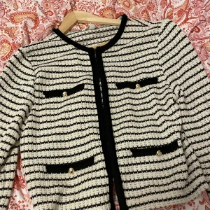 Randig stickad kofta med pärldetaljer - Säljer en snygg stickad kofta i svart och vitt med horisontella ränder. Koftan har en look som får de att se ut som du har fyra fickor framtill med pärlknappar och svarta kanter. En väldigt elegant kofta för dig som gillar klassisk men trendig stil.