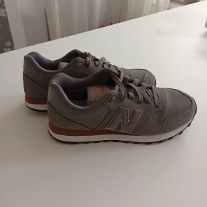 Grå New Balance sneakers - Snygga grå sneakers från New Balance med klassisk design och bruna detaljer vid sulan. Skorna har snörning, rund tå och en slitstark gummisula. Perfekta för dig som gillar en stilren och sportig look. Tillverkade i syntetmaterial och textil.