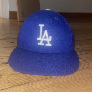 Blå LA Dodgers keps från New Era - Snygg blå keps från New Era med LA Dodgers-logga broderad framtill i vitt. Klassisk basebollmodell med böjd skärm och broderade ventilationshål. MLB-logga baktill och New Era-logga på sidan. Perfekt för dig som gillar sportig streetstyle.