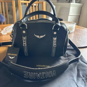 Zadig väska  - Cool svart handväska från Zadig & Voltaire i skinn med silverfärgade detaljer, dubbla handtag och avtagbar axelrem med logga. Väskan har dekorativa dragkedjor och ikonisk vinglogga framtill. Perfekt storlek för det viktigaste och ger en edgy vibe.