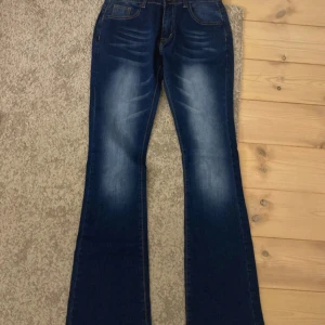 Blå bootcut jeans med utsvängda ben - Snygga blå jeans med bootcut-modell och markerade slitningar framtill. Jeansen har klassisk femficksdesign och en mörkblå tvätt med ljusare partier på låren. Perfekta för dig som gillar retrovibbar och vill ha en cool siluett. jag har aldrig använt dem men jag köpte de på Vinted och personen jag köpte de från sa att de var nya utan prislapp.