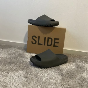 Svarta Yeezy Slides - Säljer ett par svarta Yeezy Slides med chunky sula och öppen tå. De är tillverkade i syntetmaterial och har en minimalistisk design med bred rem över foten. Perfekta för en chill och avslappnad stil.