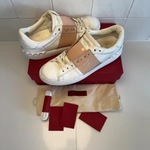 Valentino skor  - Snygga Valentino Garavani sneakers i vitt skinn med bred rosa panel och ikoniska nitar på hälen. Klassisk rund tå, platt sula och vita snören. Kommer med box!! Bara att höra av sig till oss vid minsta fundering😁