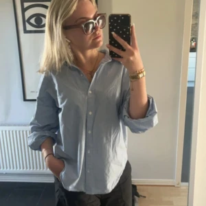 Ljusblå vit randig skjorta  - Klassisk ljusblå skjorta med knappar framtill och krage. Skjortan har lång ärm och en relaxed, lite oversized passform. Perfekt att styla med smycken och uppkavlade ärmar för en chill vibe.