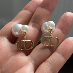 Dior örhängen med pärlor och guldlogo - Snygga örhängen från Dior med dubbla vita pärlor och hängande guldfärgade CD-loggor. Perfekt statement-accessoar som lyfter vilken outfit som helst. Klassisk design med lyxig känsla och trendig touch.