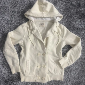 Vit hoodie-jacka från Big Star - Mysig vit hoodie-jacka från Big Star med dubbelknäppning framtill och stora knappar. Jackan har huva med dragsko, två fickor och är fodrad med mjuk fleece. Perfekt för höst dagar och har en clean, stilren look.