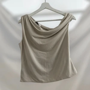 Beige omlott ärmlös topp - Snygg beige topp med draperad halsringning och omlottkänsla. Ärmlös design med en axel lätt rynkad för extra detalj. Perfekt för dig som gillar stilrena och trendiga plagg med en twist.
