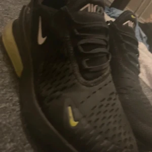 Nike Air Max 270 svarta sneakers - Svarta Nike Air Max 270 sneakers med gula detaljer och vit logga. Ovandelen är i ventilerande syntetmaterial med snörning och rund tå. Sulan har en tydlig air-enhet baktill och gult inslag på sidan. Perfekta för dig som gillar sportig och modern stil. Lite färg fläckar som jag tyvärr inte tror att man kan få bort men är inte säker har bara försökt med vatten och ett hål vid tån