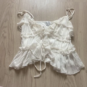 Vit plisserad linnetopp med volanger - Supersöt vit linnetopp från Monki med plisserat tyg, volangdetaljer och knyt framtill. Axelbanden är smala och toppen har en luftig, flowy känsla som passar perfekt till varma dagar.