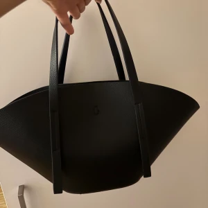 Svart shoppingväska i skinn - Stilren svart shoppingväska med unik, bred form och långa handtag. Väskan är tillverkad i slätt skinn och har en minimalistisk design utan synliga loggor eller detaljer. Perfekt för dig som gillar en clean look och vill ha plats för allt du behöver under dagen.