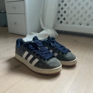 mörkblå Adidas Campus  - Snygga Adidas Campus sneakers i mörkblå mocka med klassiska vita ränder på sidorna och mörkblå snören. Skorna har en beige sula och Adidas logga på hälen. 